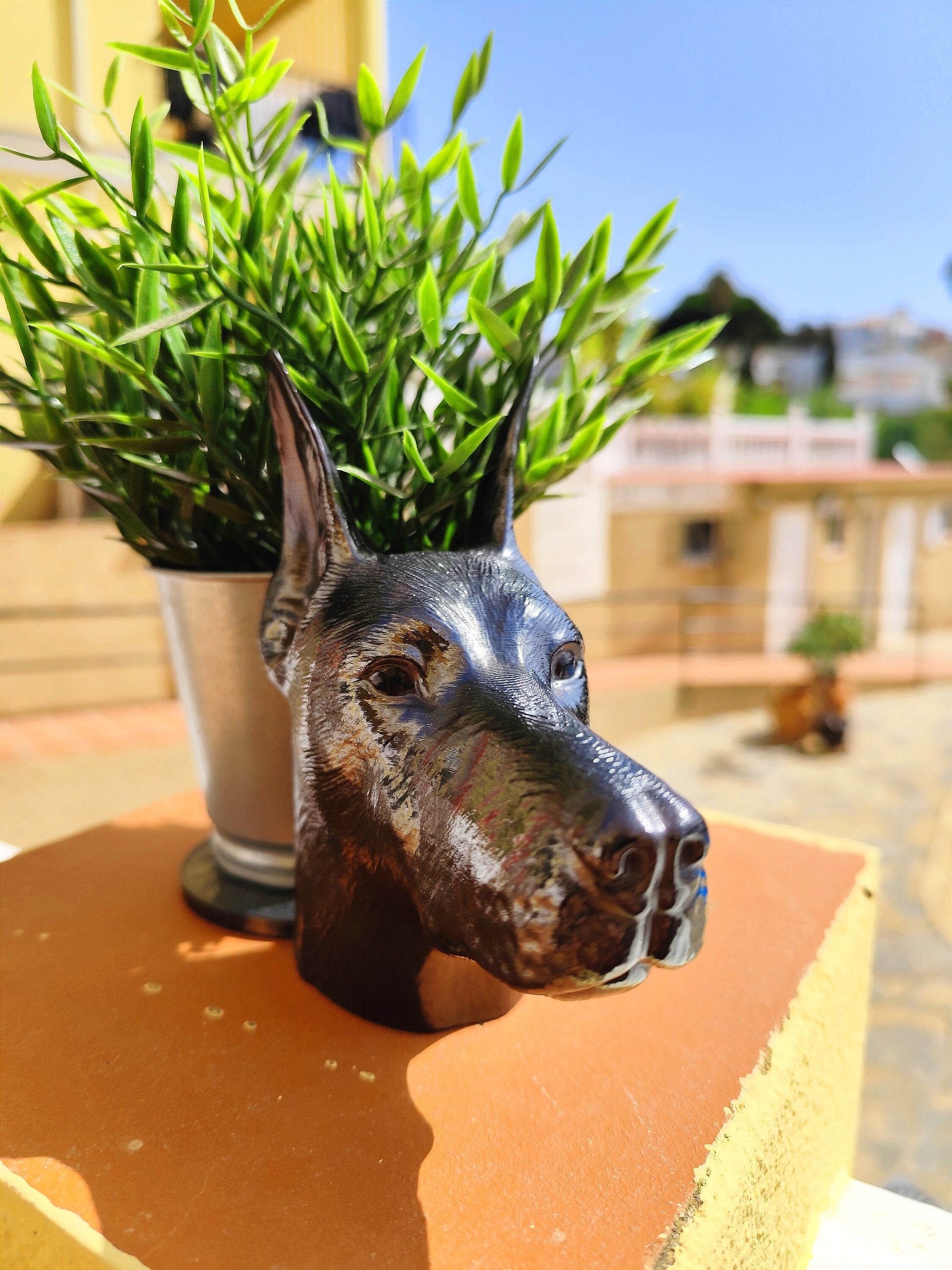 Side view of a hand-painted Doberman planter with metallic gold and silver accents, showcasing the Doberman&#39;s sleek profile. A green plant is placed inside the planter, creating a stylish décor piece for indoor or outdoor use.