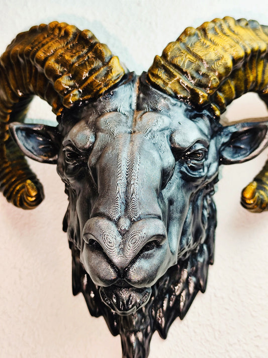 Front view of a hand-painted ram head wall mount with metallic gold and silver detailing. The sculpture features intricately designed horns and lifelike facial details, mounted on a white textured wall