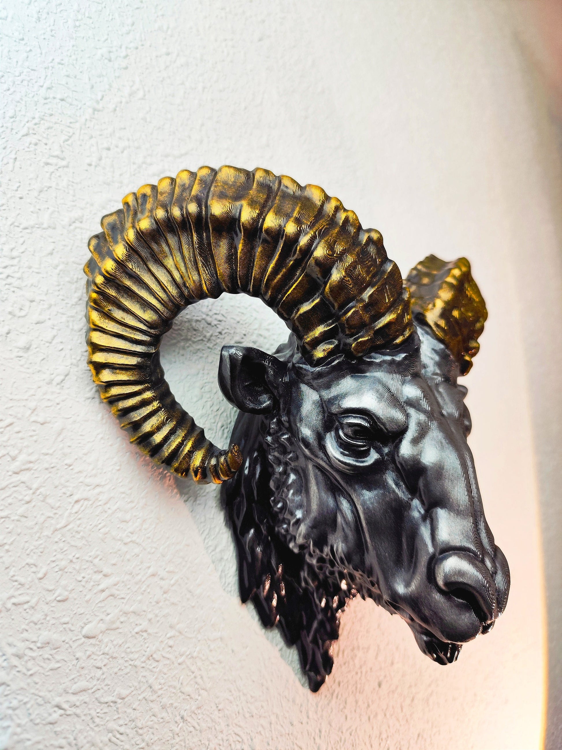 Side view of a hand-painted ram head wall mount, showcasing the intricate metallic gold and silver details on the horns and realistic facial structure. The sculpture is mounted on a white wall, highlighting its striking design