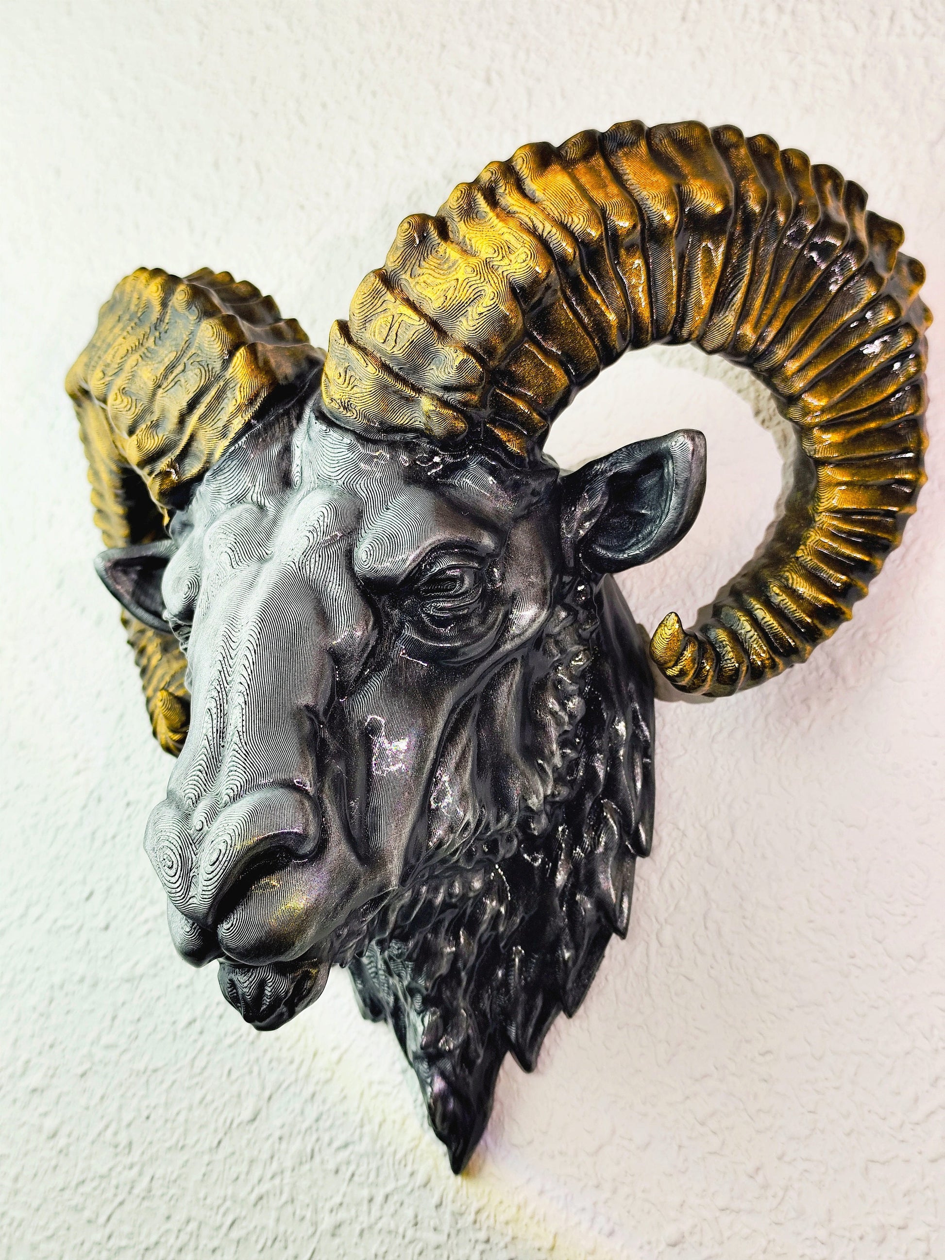 Side view of a hand-painted ram head wall mount, showcasing the intricate metallic gold and silver details on the horns and realistic facial structure. The sculpture is mounted on a white wall, highlighting its striking design