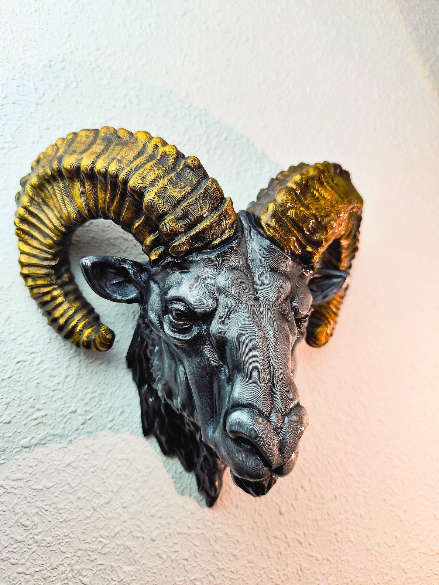 Side view of a hand-painted ram head wall mount, showcasing the intricate metallic gold and silver details on the horns and realistic facial structure. The sculpture is mounted on a white wall, highlighting its striking design