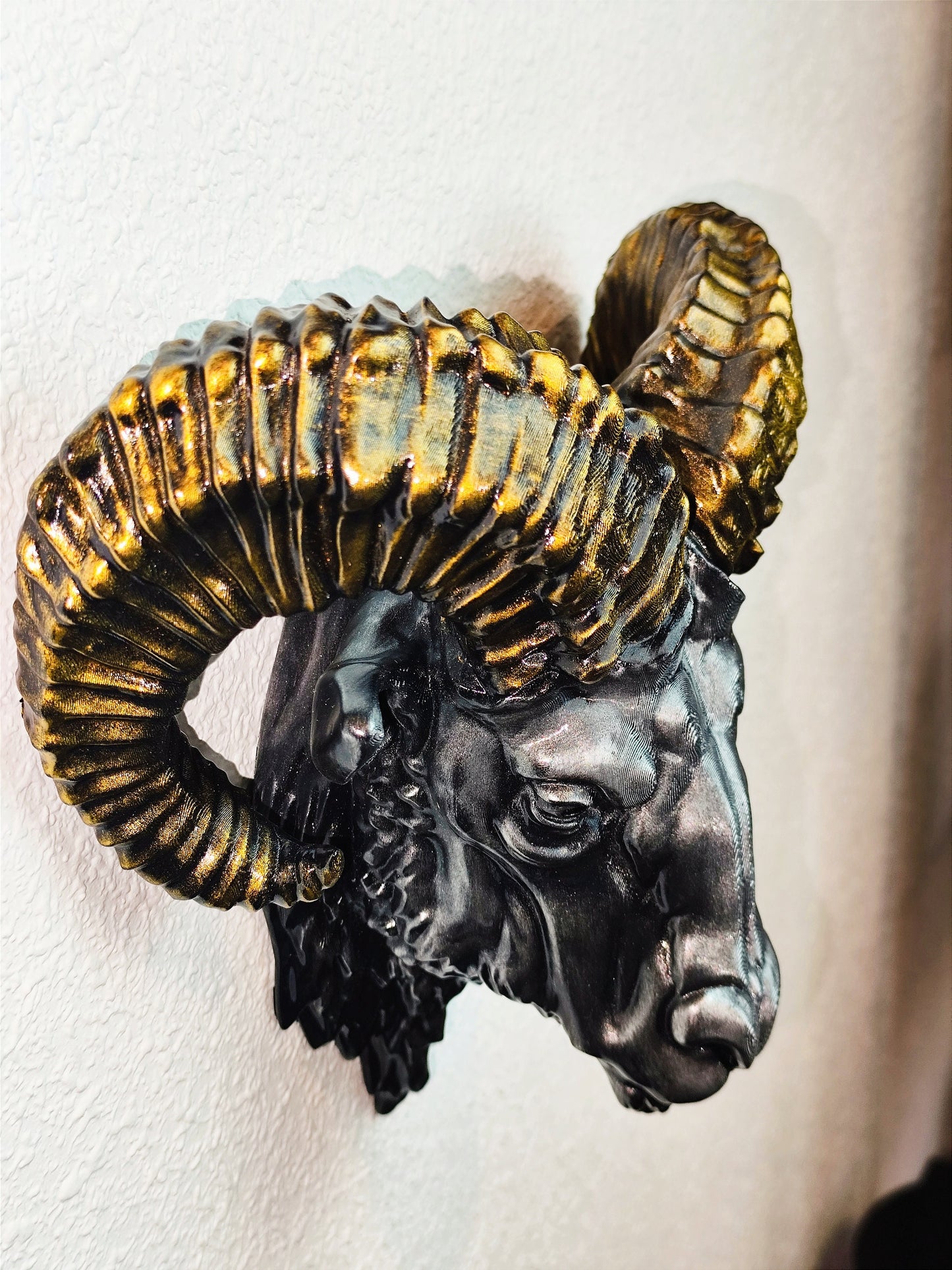 Side view of a hand-painted ram head wall mount, showcasing the intricate metallic gold and silver details on the horns and realistic facial structure. The sculpture is mounted on a white wall, highlighting its striking design