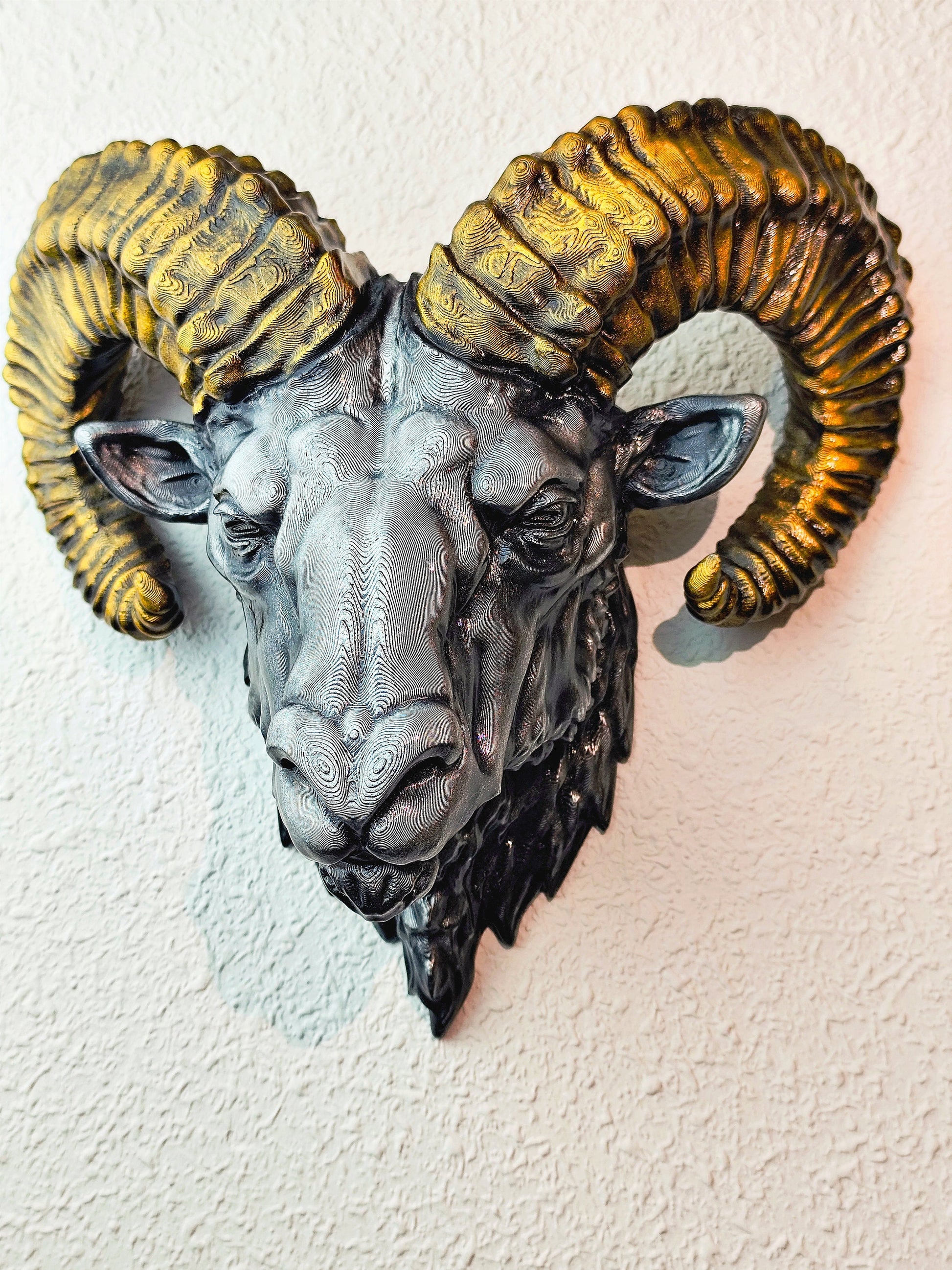 Side view of a hand-painted ram head wall mount, showcasing the intricate metallic gold and silver details on the horns and realistic facial structure. The sculpture is mounted on a white wall, highlighting its striking design