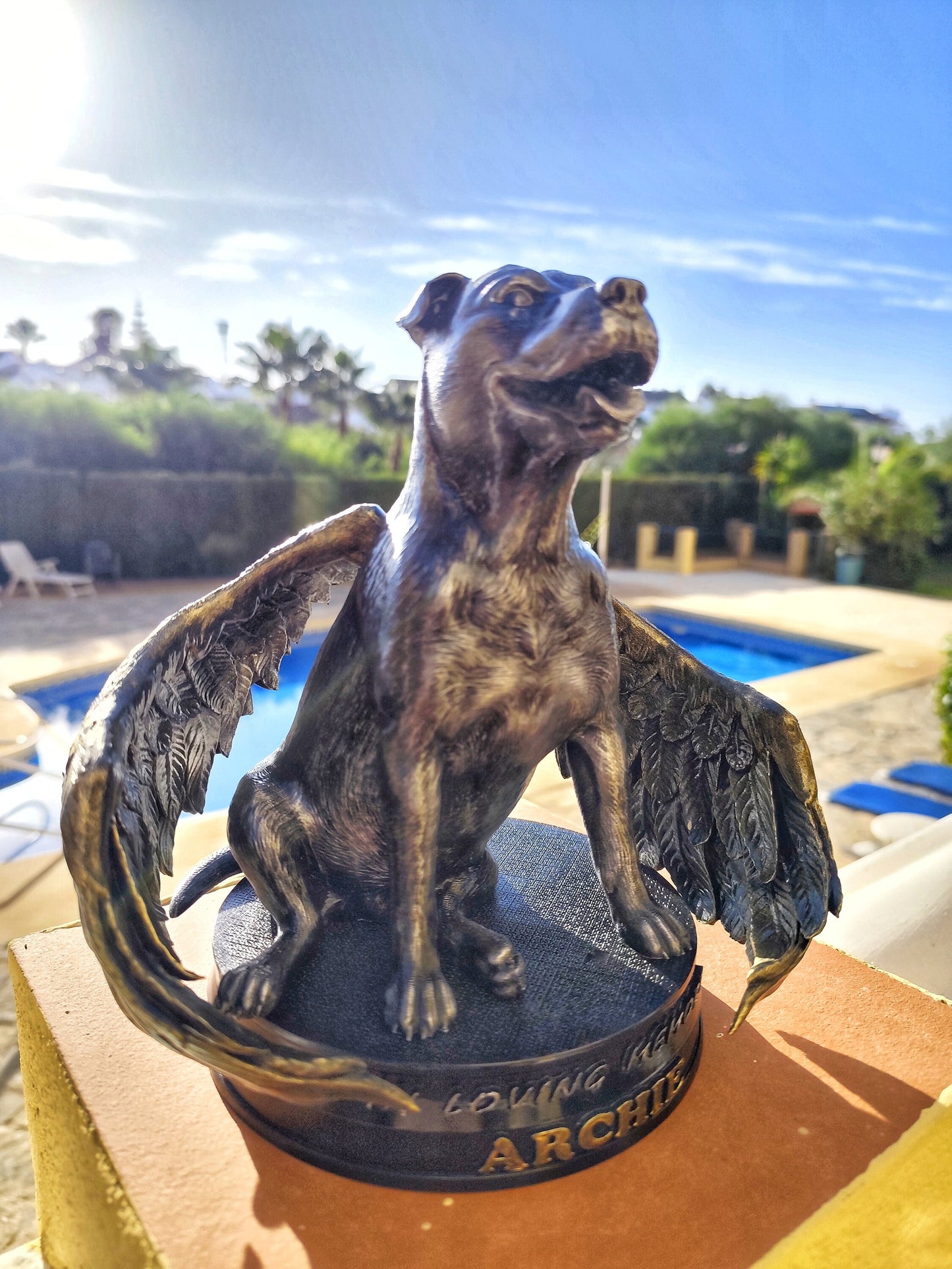 A memorial statue of a smiling dog with angel wings, resembling a pit bull breed, standing on a circular base. The wings are large, detailed with golden feathers