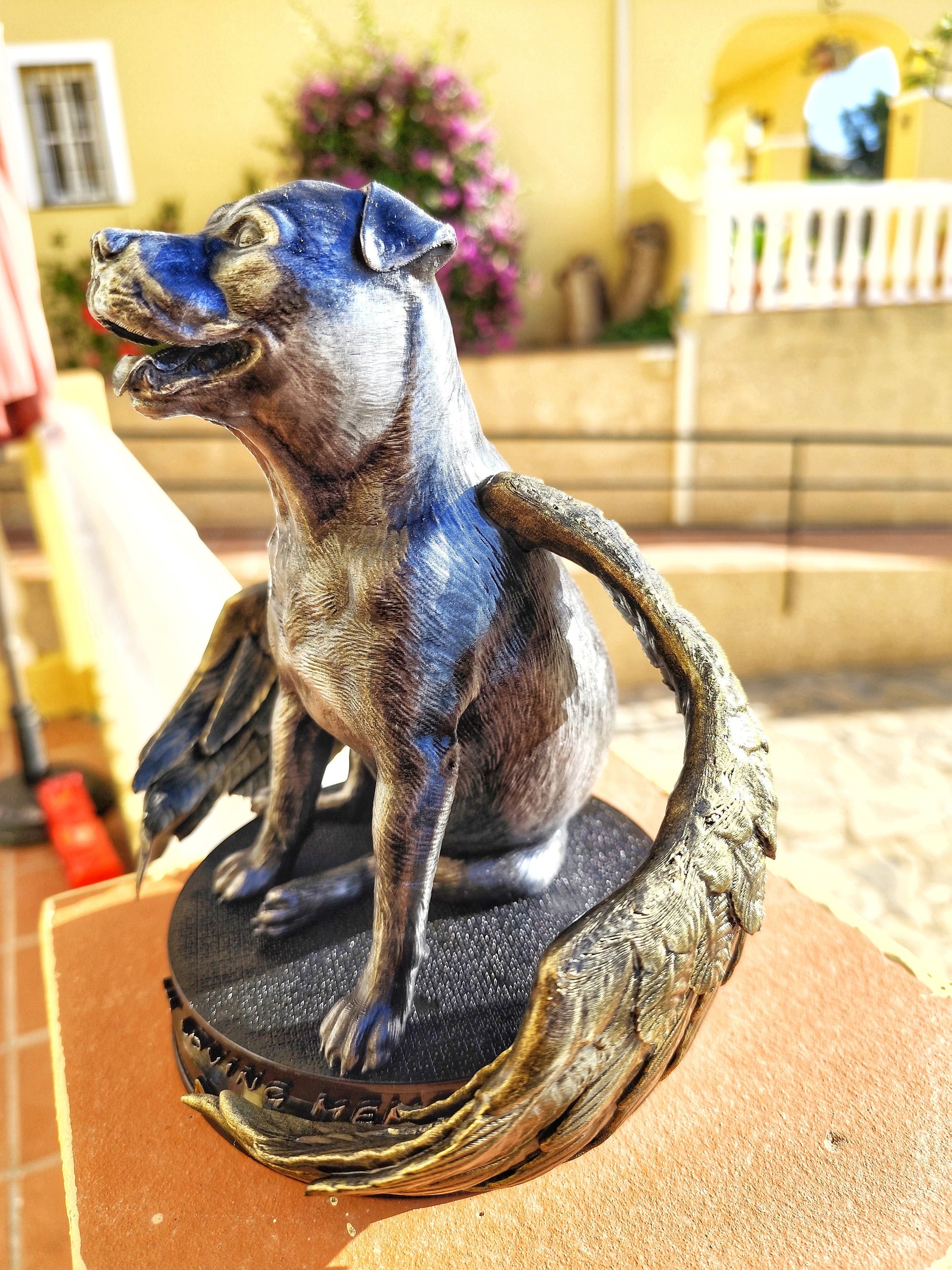 A memorial statue of a smiling dog with angel wings, resembling a pit bull breed, standing on a circular base. The wings are large, detailed with golden feathers