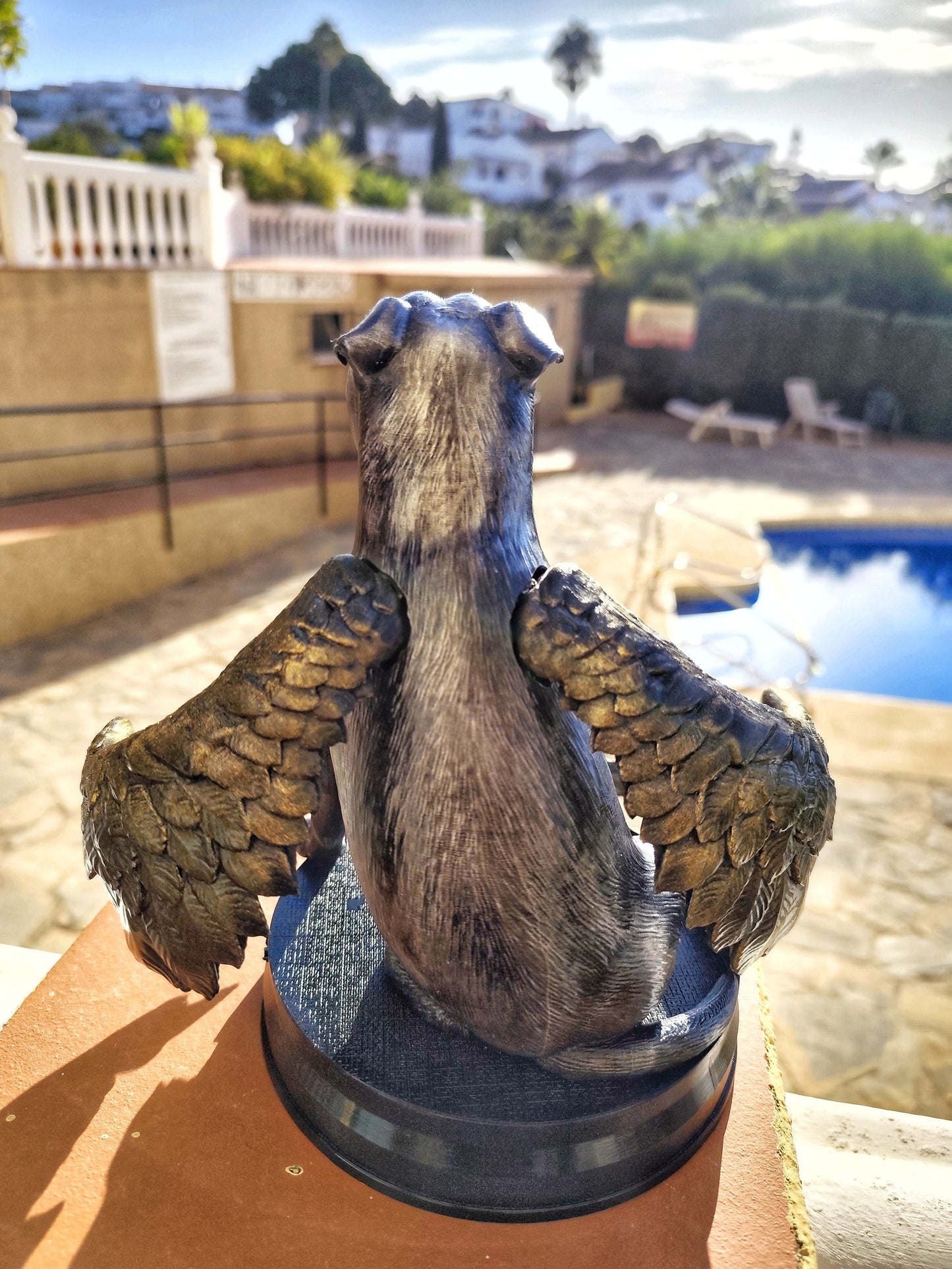 A memorial statue of a smiling dog with angel wings, resembling a pit bull breed, standing on a circular base. The wings are large, detailed with golden feathers