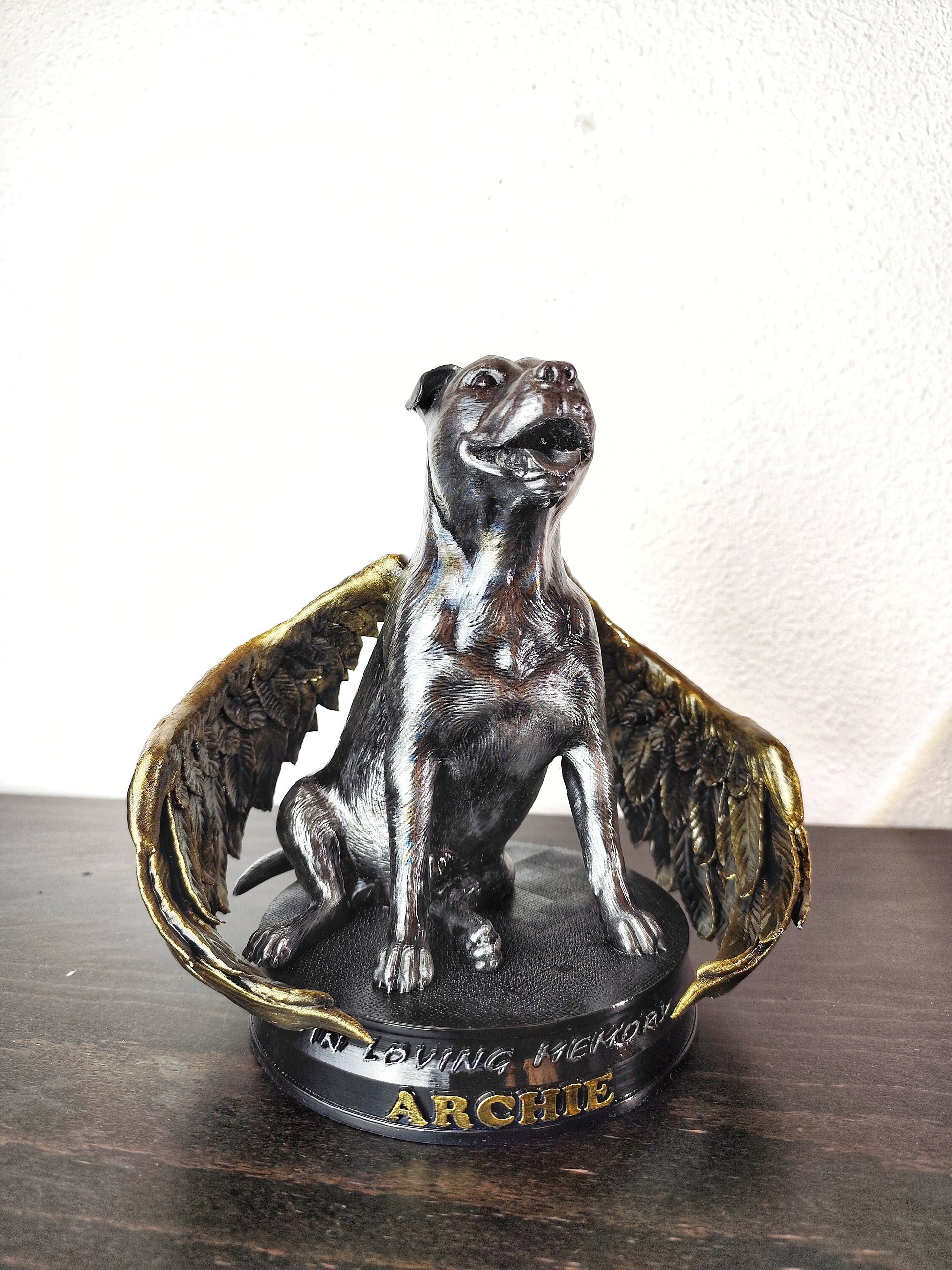 A memorial statue of a smiling dog with angel wings, resembling a pit bull breed, standing on a circular base. The wings are large, detailed with golden feathers