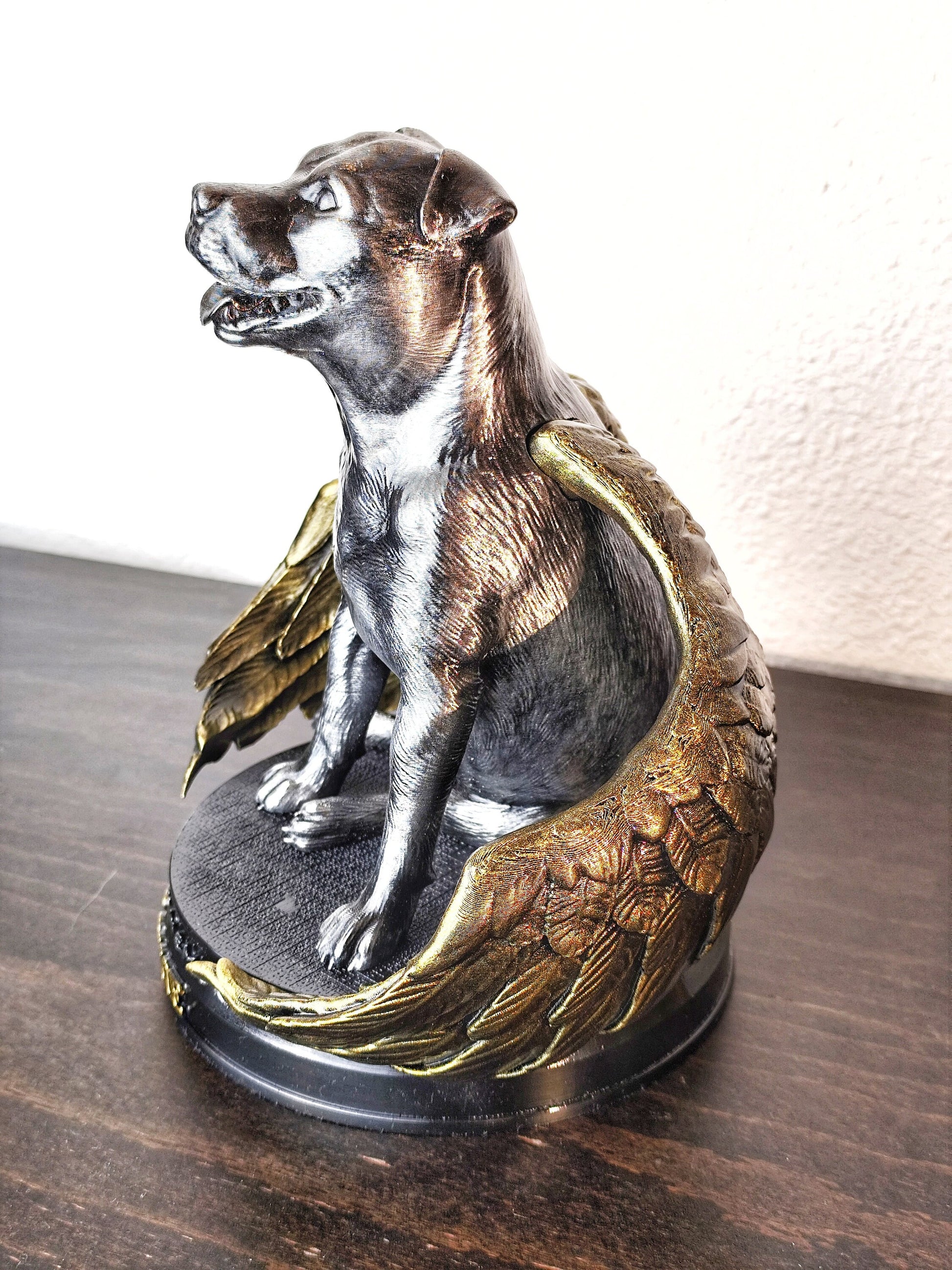 A memorial statue of a smiling dog with angel wings, resembling a pit bull breed, standing on a circular base. The wings are large, detailed with golden feathers