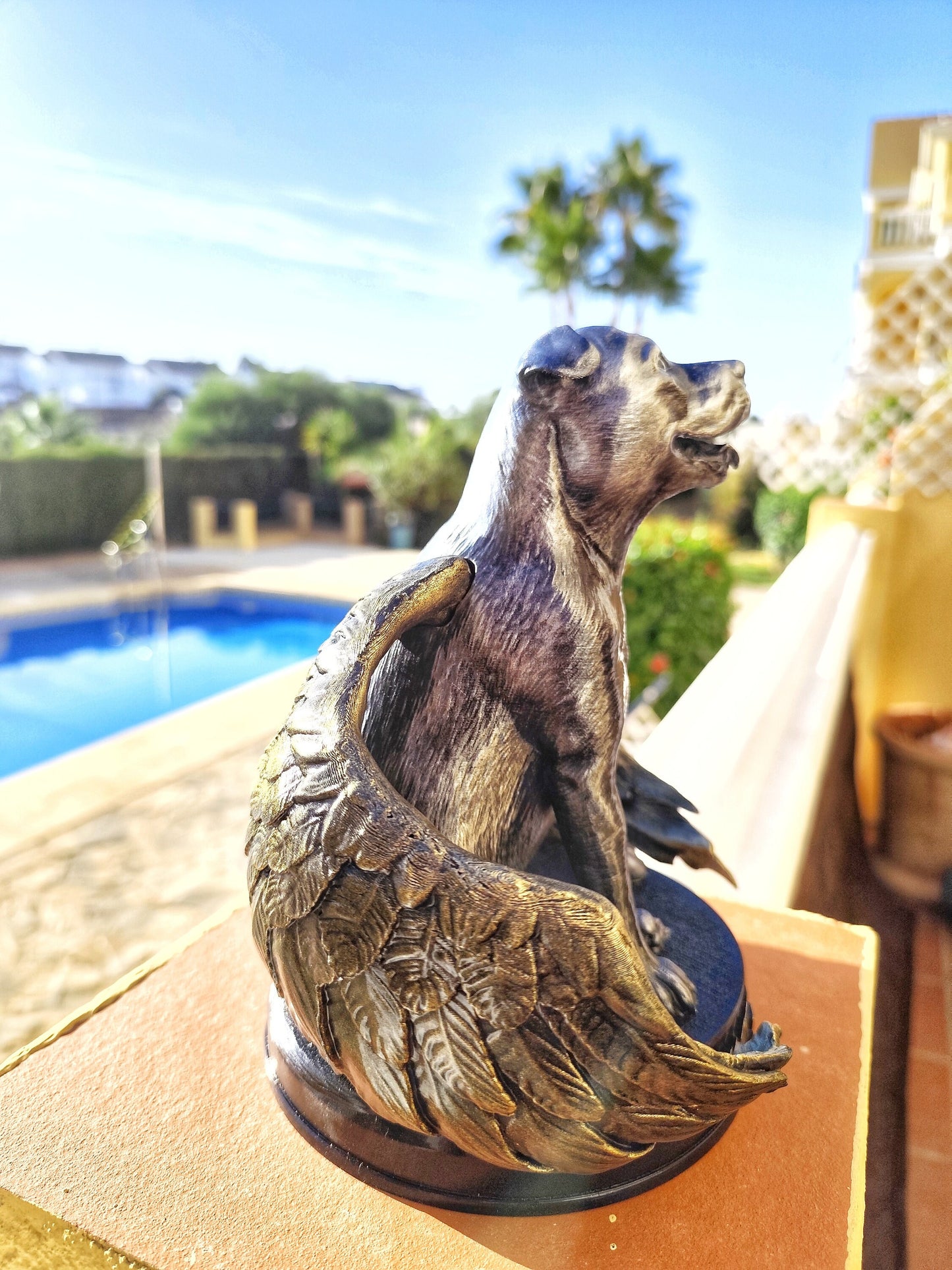 A memorial statue of a smiling dog with angel wings, resembling a pit bull breed, standing on a circular base. The wings are large, detailed with golden feathers