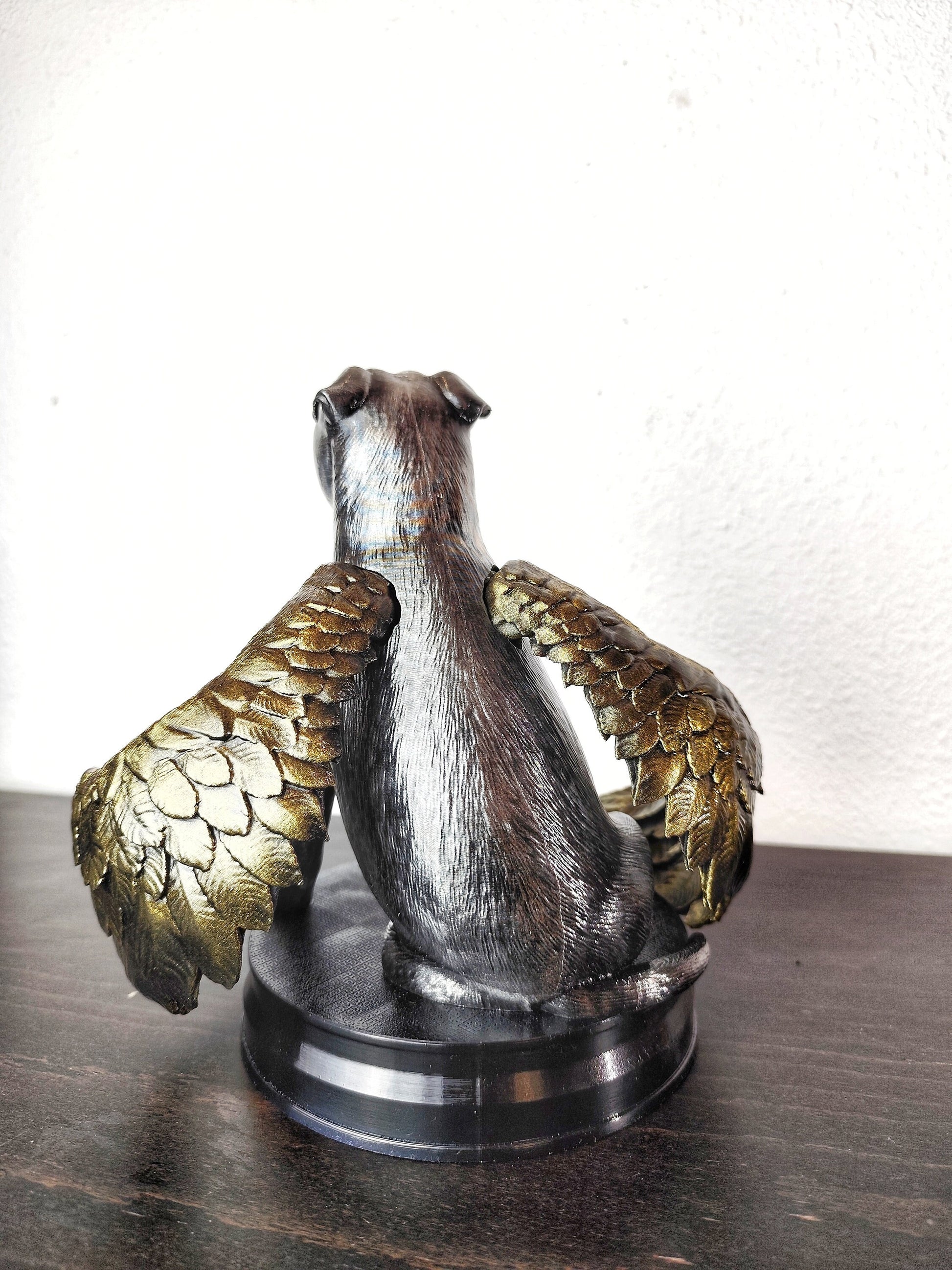 A memorial statue of a smiling dog with angel wings, resembling a pit bull breed, standing on a circular base. The wings are large, detailed with golden feathers