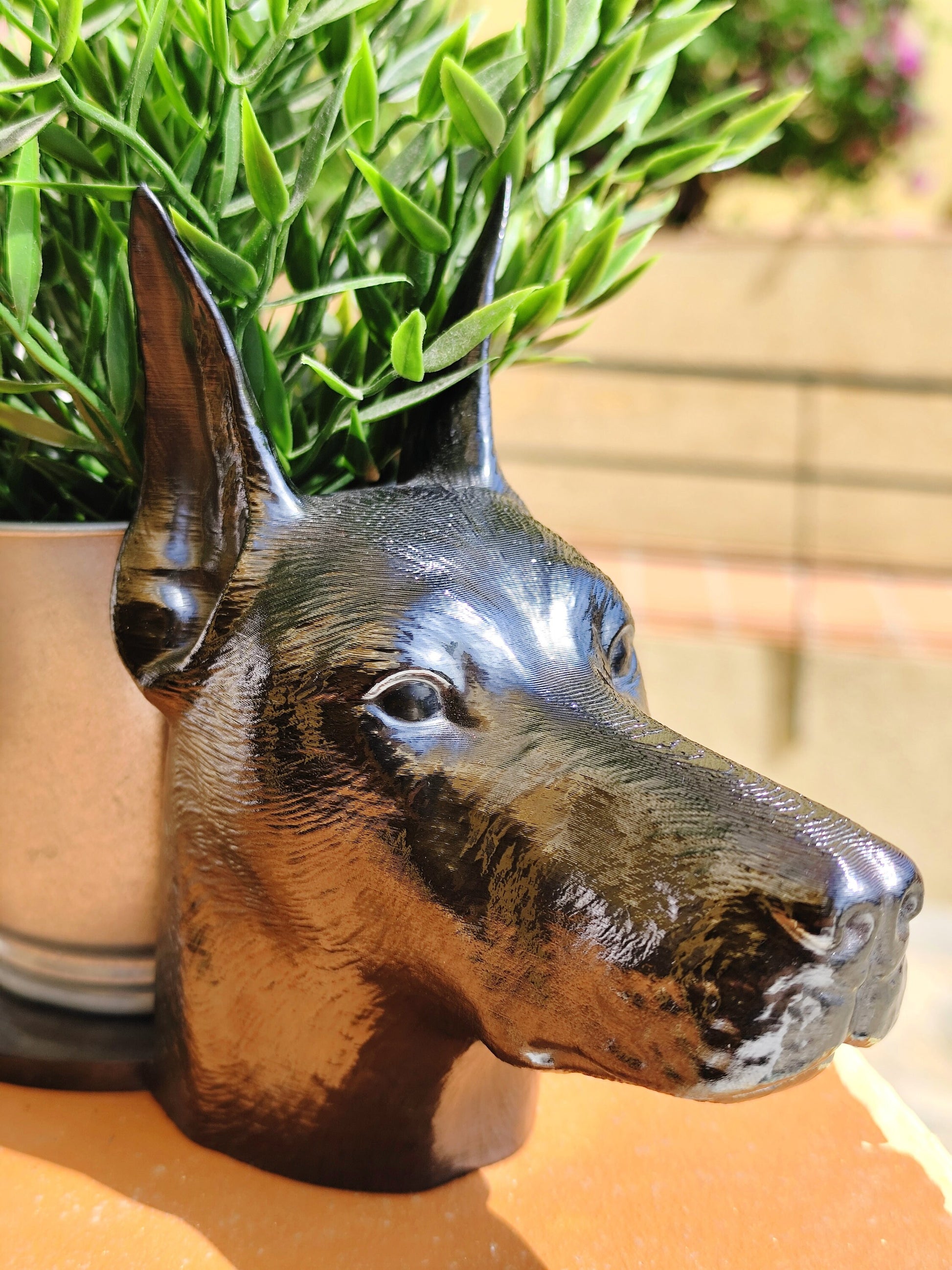 Side view of a hand-painted Doberman planter with metallic gold and silver accents, showcasing the Doberman&#39;s sleek profile. A green plant is placed inside the planter, creating a stylish décor piece for indoor or outdoor use.