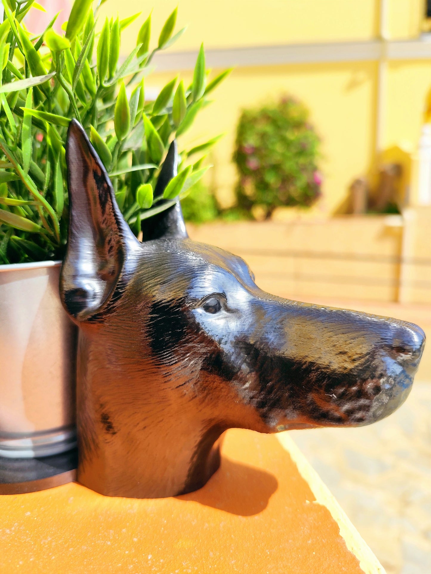 Side view of a hand-painted Doberman planter with metallic gold and silver accents, showcasing the Doberman&#39;s sleek profile. A green plant is placed inside the planter, creating a stylish décor piece for indoor or outdoor use.