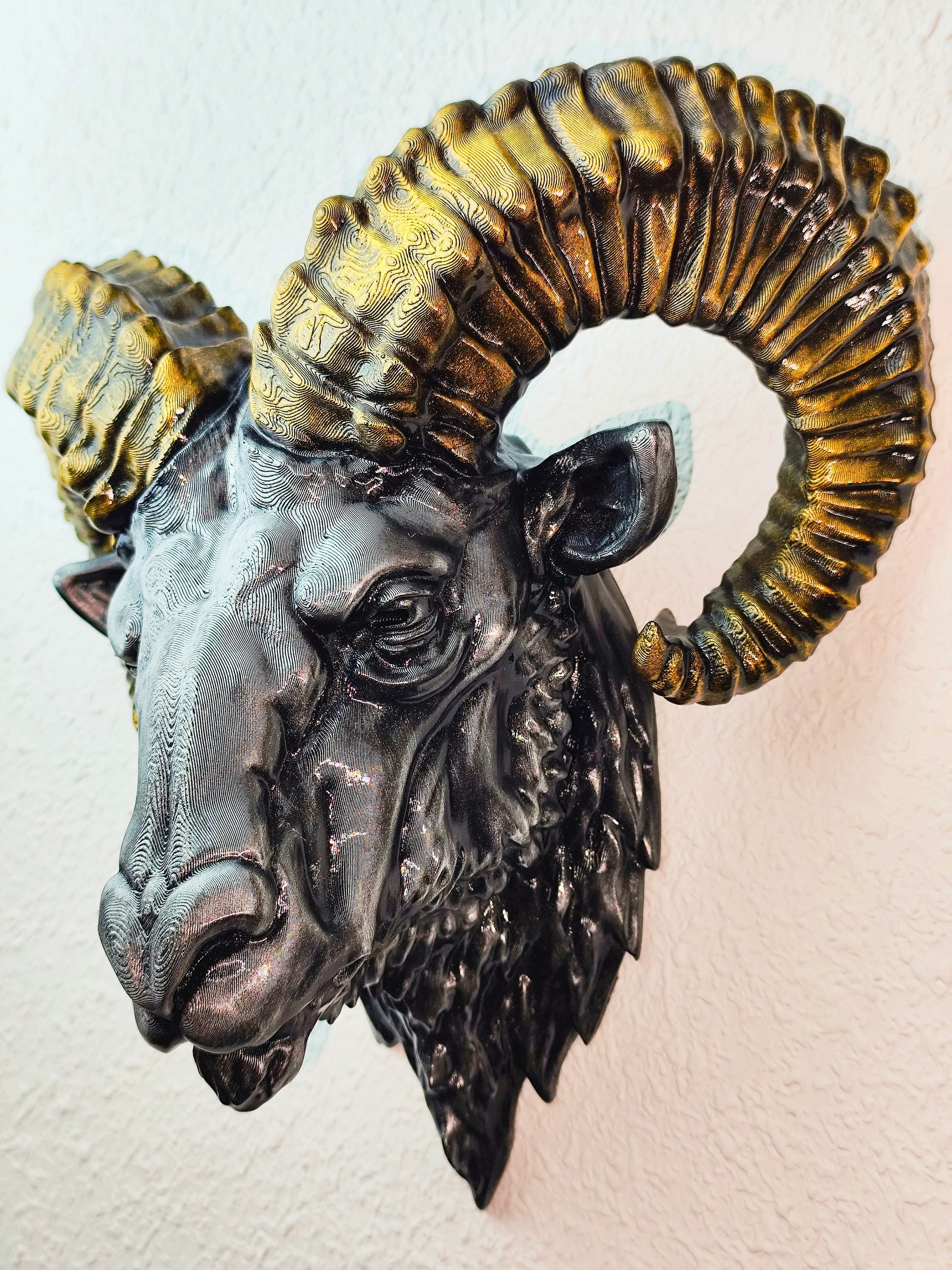 Side view of a hand-painted ram head wall mount, showcasing the intricate metallic gold and silver details on the horns and realistic facial structure. The sculpture is mounted on a white wall, highlighting its striking design