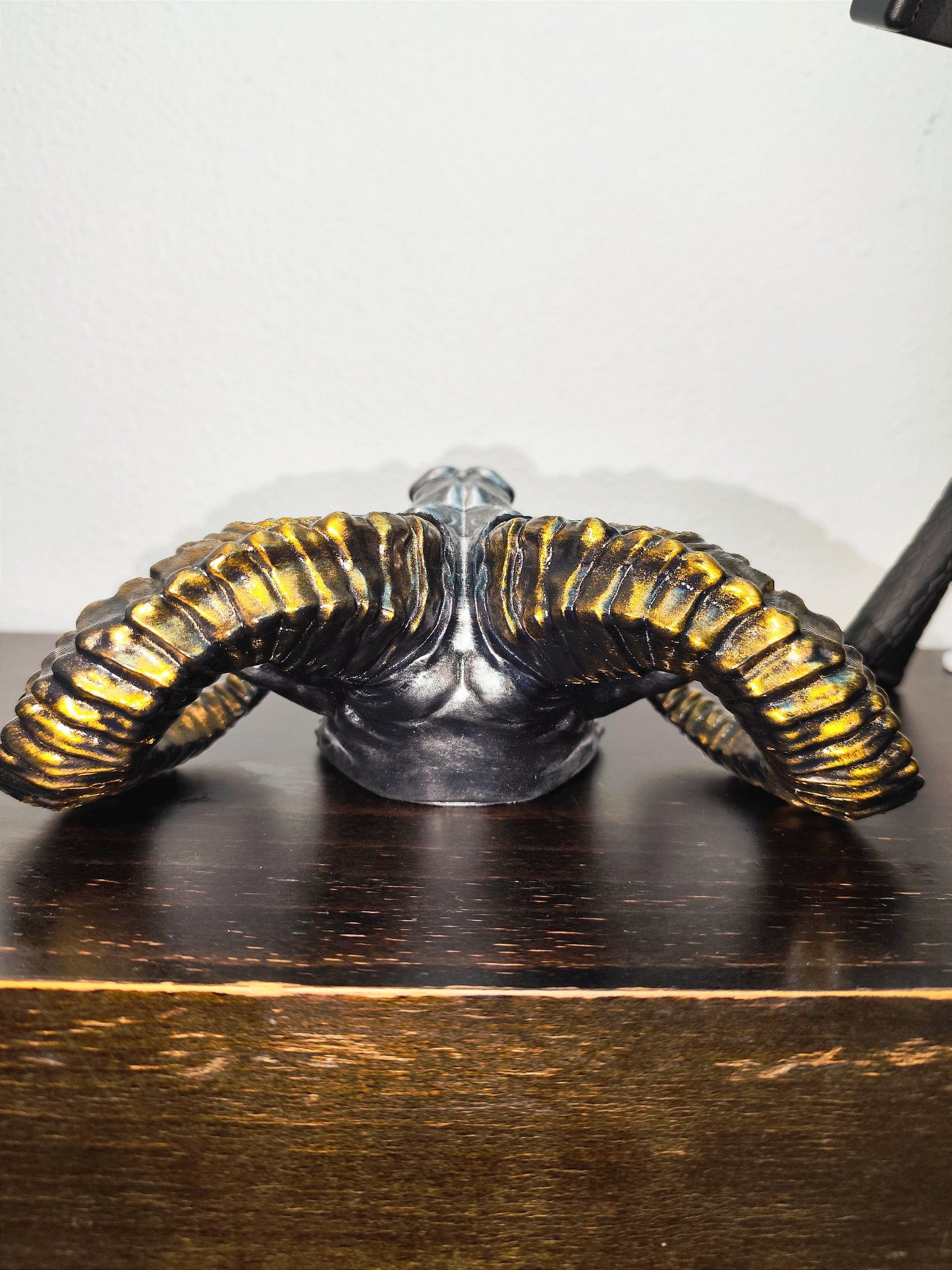 Side view of a hand-painted ram head wall mount, showcasing the intricate metallic gold and silver details on the horns and realistic facial structure. The sculpture is mounted on a white wall, highlighting its striking design