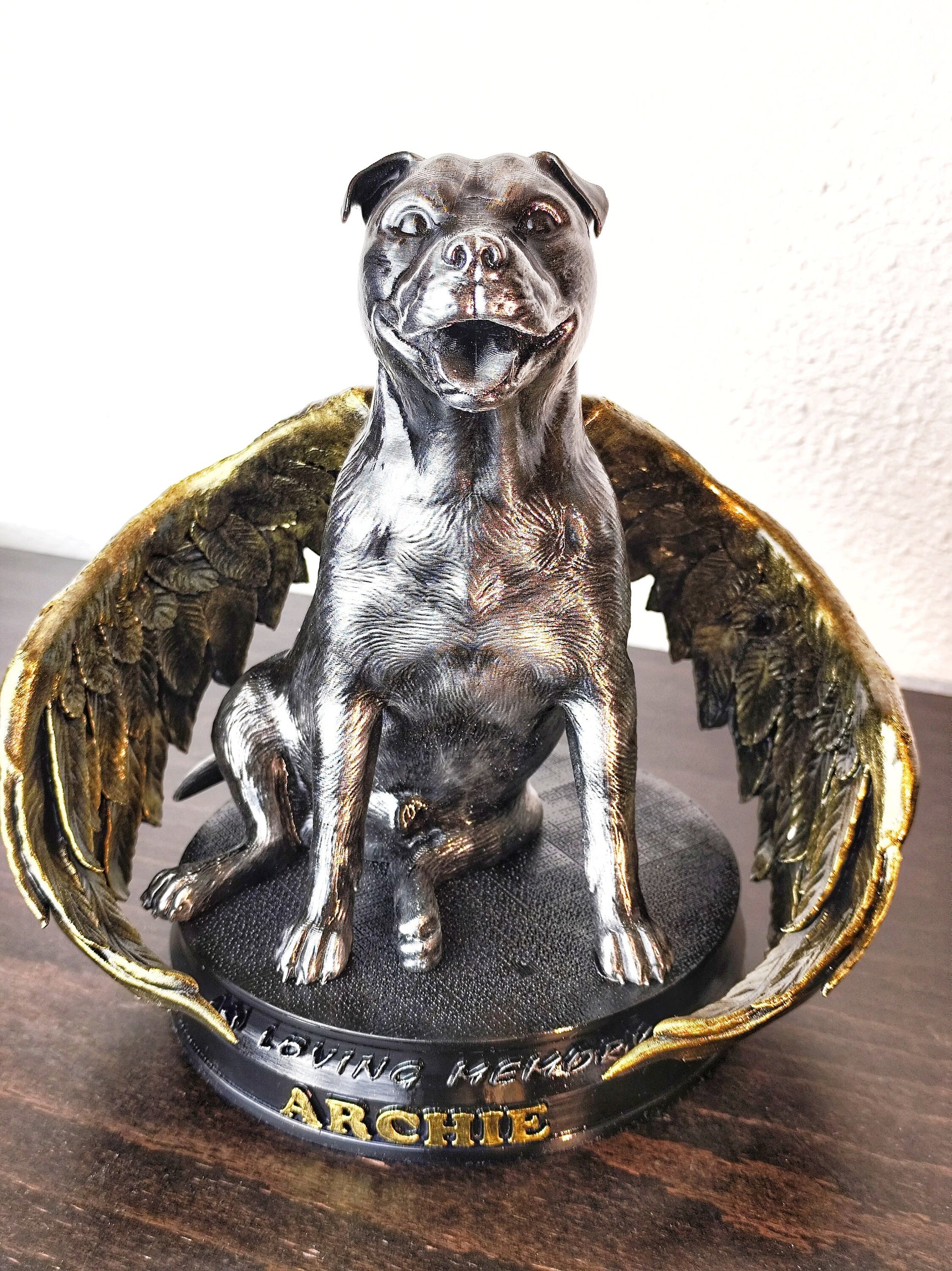 A side view of a memorial dog statue with angel wings, resembling a pit bull. The statue sits on a circular base with gold feathered wings curving around the dog&#39;s body. The dog has a joyful expression and the metallic, bronze-colored surface shines