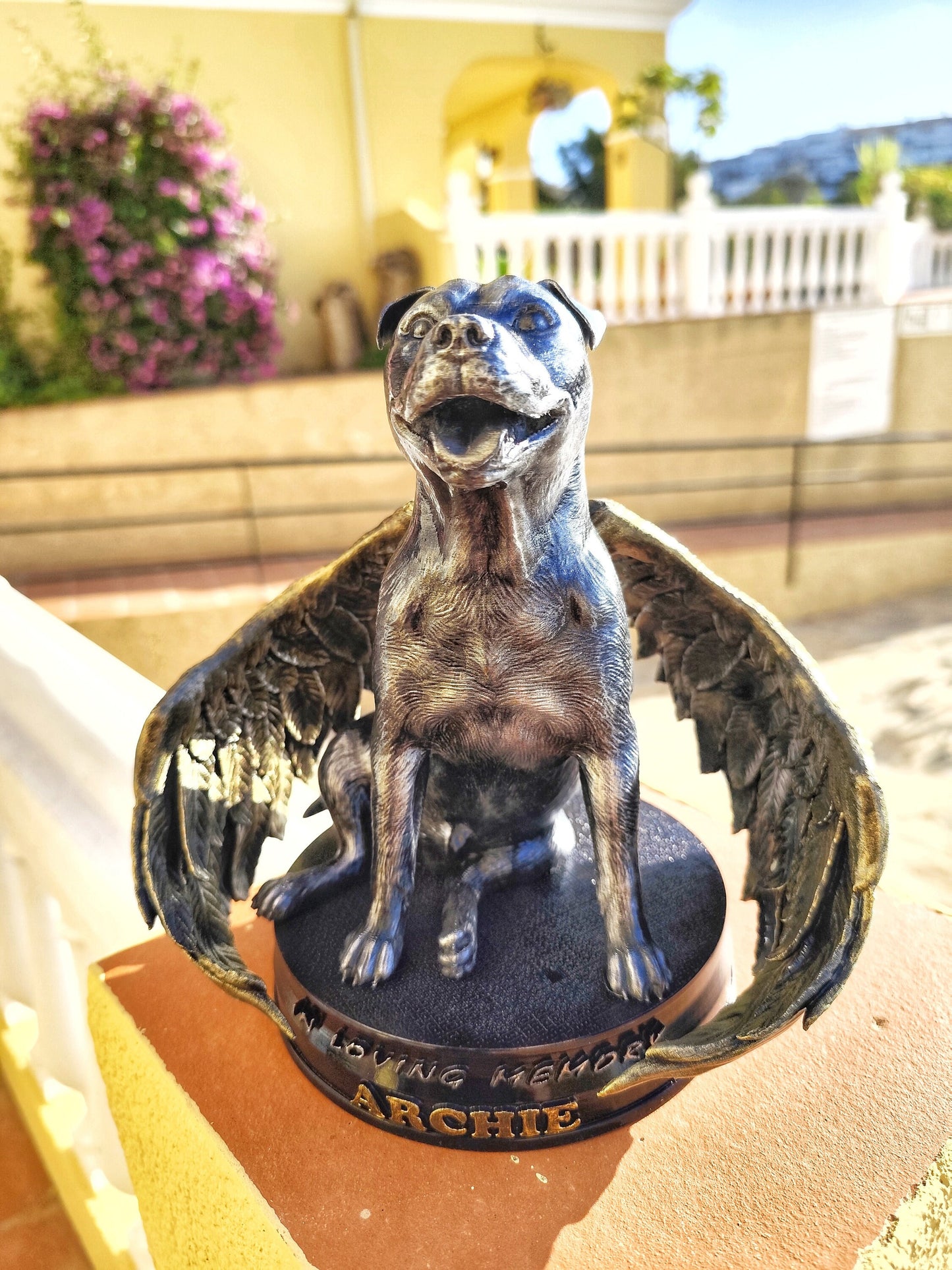 A memorial statue of a smiling dog with angel wings, resembling a Staffordshire bull breed, standing on a circular base. The wings are large, detailed with golden feathers